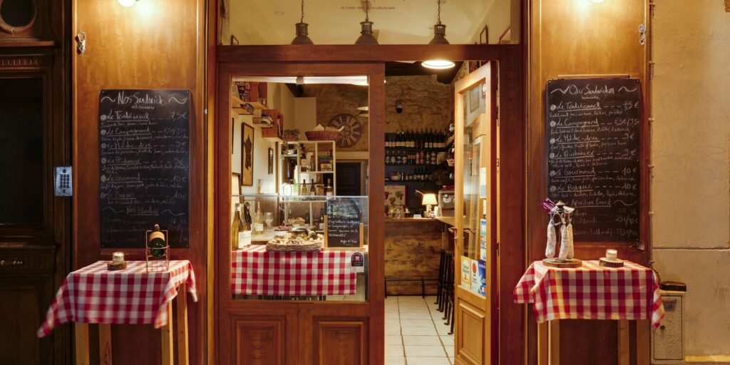 le forum du rotary une petite entrée de restaurant avec des portes en bois vous invite à une balade historique, avec des menus sur tableau noir, des nappes à carreaux rouges et blancs, et un intérieur bien éclairé avec des étagères de vins et de produits gastronomiques.
