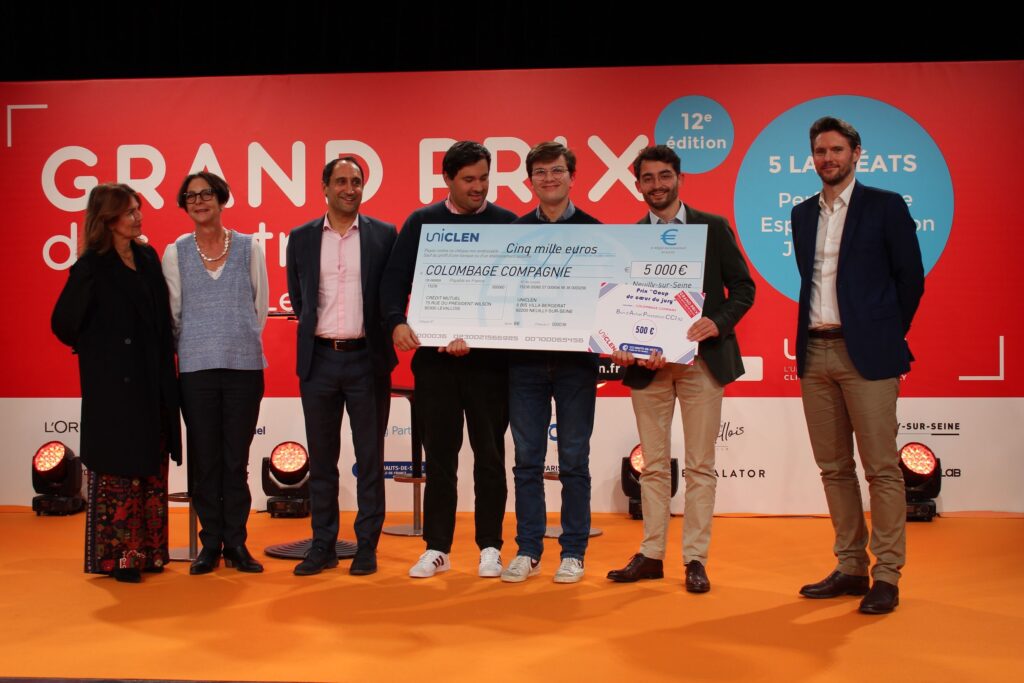 le forum du rotary un groupe de sept personnes se tient sur une scène orange. au centre, trois hommes, reconnus pour leur talent, tiennent un gros chèque de 5 000 euros sur lequel est inscrit "colombage compagnie". une grande banderole rouge du grand prix se trouve à l'arrière plan.