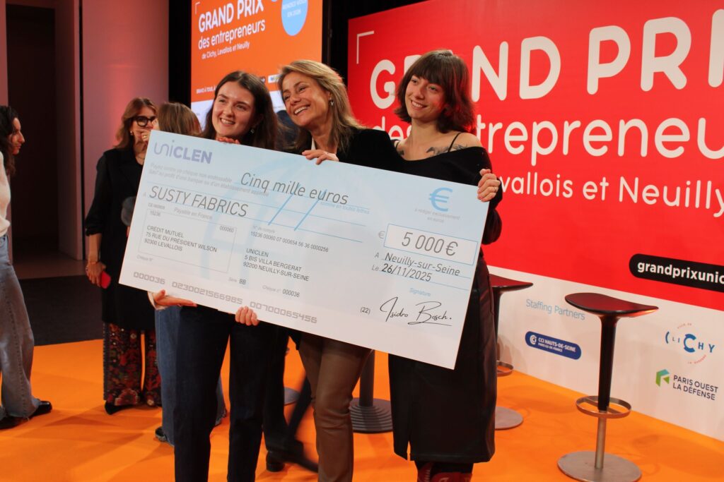 le forum du rotary trois femmes talentueuses se tiennent sur une scène orange, tenant un gros chèque de 5 000 euros à l'ordre de susty fabrics, à l'occasion du grand prix des entrepreneurs, qui célèbre l'innovation dans les start ups. une bannière rouge et blanche de l'événement est visible derrière elles.