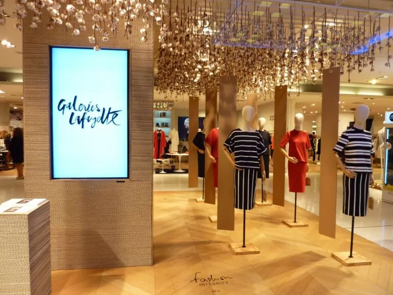 le forum du rotary l'étalage d'un magasin de vêtements avec des mannequins vêtus de robes rayées et unies illustre l'essence de la mode française. un grand écran affiche "galeries lafayette", avec en arrière plan des éléments décoratifs suspendus, des clients et des étagères de vêtements.