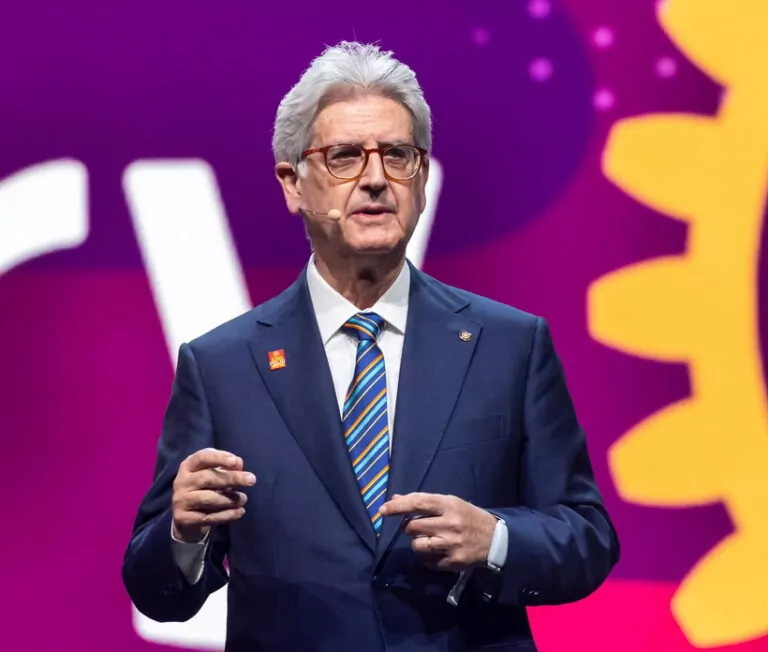 Le Forum du Rotary - Un homme en costume bleu foncé, cravate rayée et lunettes s'exprime sur scène lors d'un forum rotarien à l'aide d'un micro-casque. L'arrière-plan est violet avec des formes abstraites jaunes et blanches.