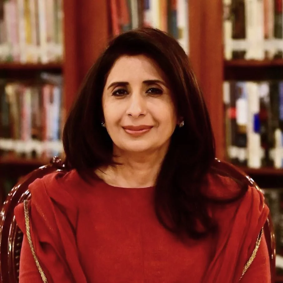 le forum du rotary mumtaz zahra baloch, une femme aux cheveux noirs portant un vêtement rouge, est assise sur une chaise devant des étagères remplies de livres, regardant la caméra et souriant légèrement.