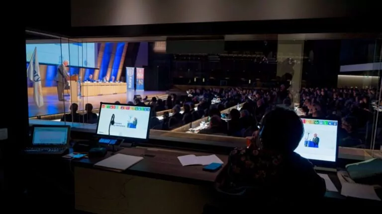 le forum du rotary vue d'une cabine de traduction donnant sur une salle de conférence, avec une personne à un bureau surveillant des écrans et le public regardant un orateur à la tribune. divers écrans, documents et informations pratiques sont visibles dans la cabine.
