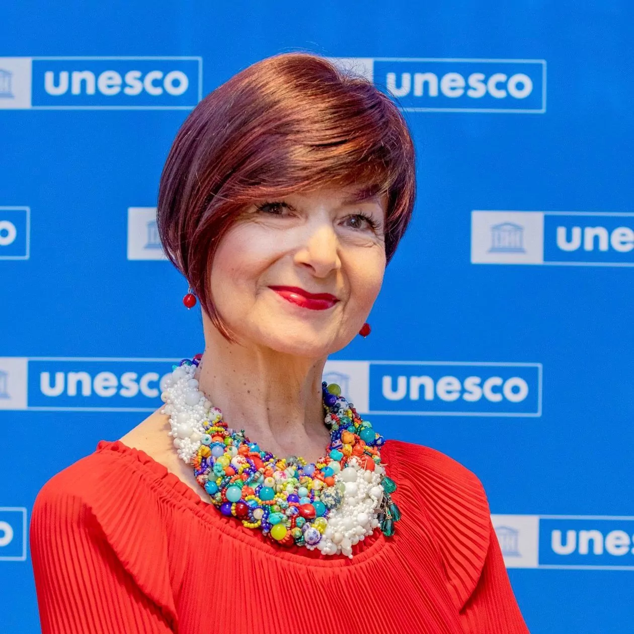 le forum du rotary s.e. simona mirela miculescu, cheveux courts rouge foncé et collier de perles coloré, se tient devant un fond bleu orné de multiples logos de l'unesco. elle porte un haut rouge et sourit chaleureusement à la caméra.