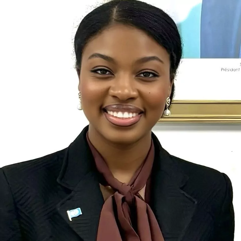 le forum du rotary syndie amoussou fléan, vêtue d'un blazer sombre, d'un chemisier marron avec un nœud papillon, de boucles d'oreilles en perles et d'une broche, sourit à la caméra. une photo encadrée est accrochée au mur derrière elle.