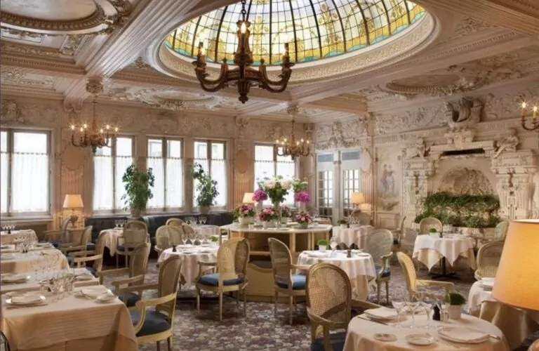 le forum du rotary une élégante salle à manger de restaurant avec un décor orné, des lustres, un grand plafond en verre teinté, des tables rondes recouvertes de nappes blanches, des chaises rembourrées, des centres de table floraux et des sculptures murales décoratives.