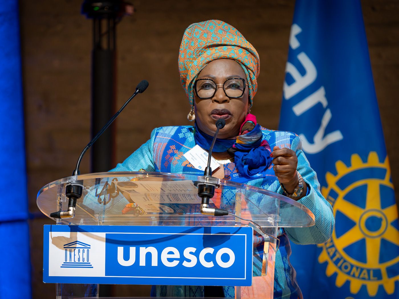 le forum du rotary une femme portant un bandeau à motifs et des lunettes parle sur un podium clair avec un signe de l'unesco. elle tient un stylo et du papier. l'arrière plan comprend un drapeau de l'unesco et une partie du logo de la roue du rotary international.