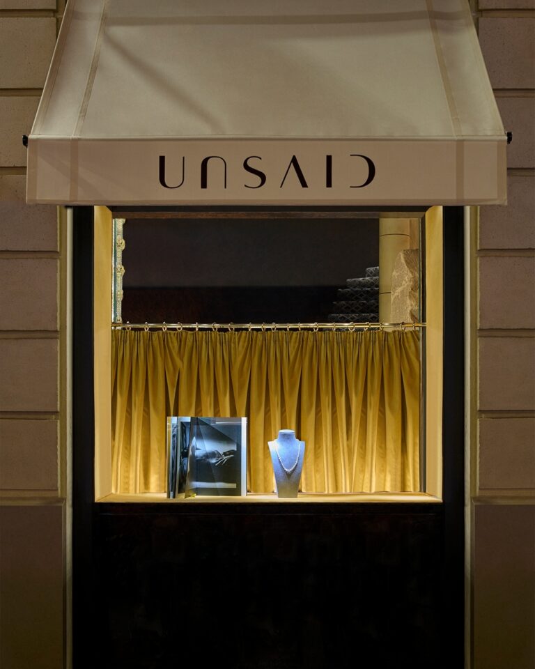 le forum du rotary vitrine d'une bijouterie avec un auvent crème portant l'inscription "unsaid" à l'envers, des rideaux jaunes, un collier éclairé en bleu sur un support et un livre ouvert posé à côté.