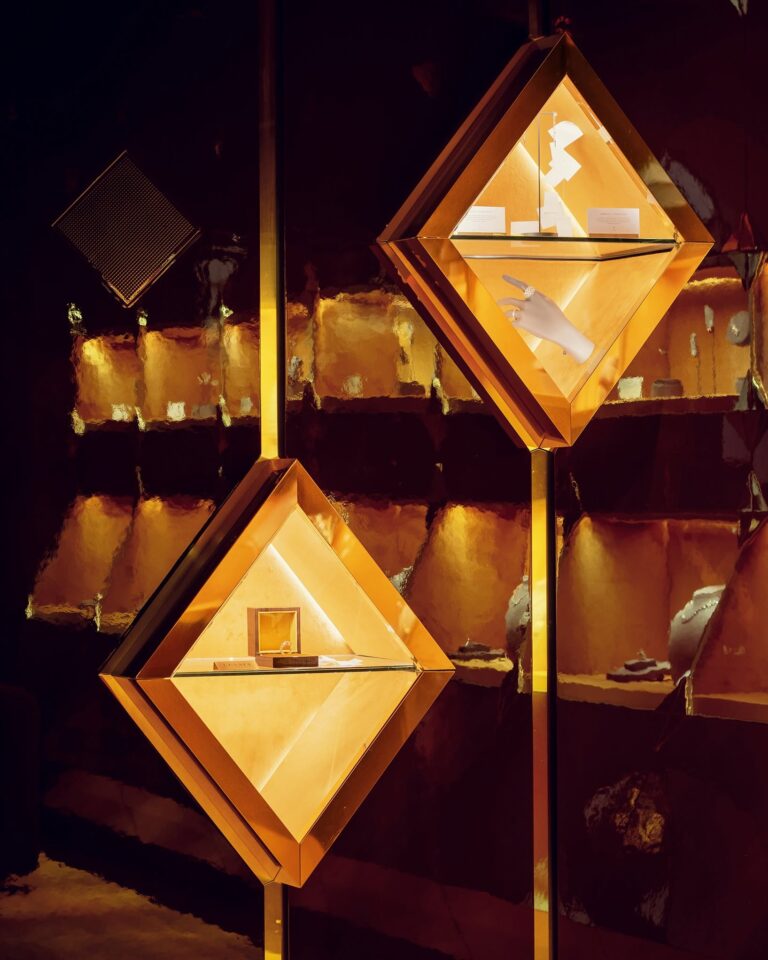 le forum du rotary deux vitrines éclairées, en forme de losange, montées sur un mur. les vitrines contiennent une sculpture blanche, un gant blanc et de petites affiches. l'arrière plan est d'un ton chaud, brun doré, avec des étagères et un éclairage doux.