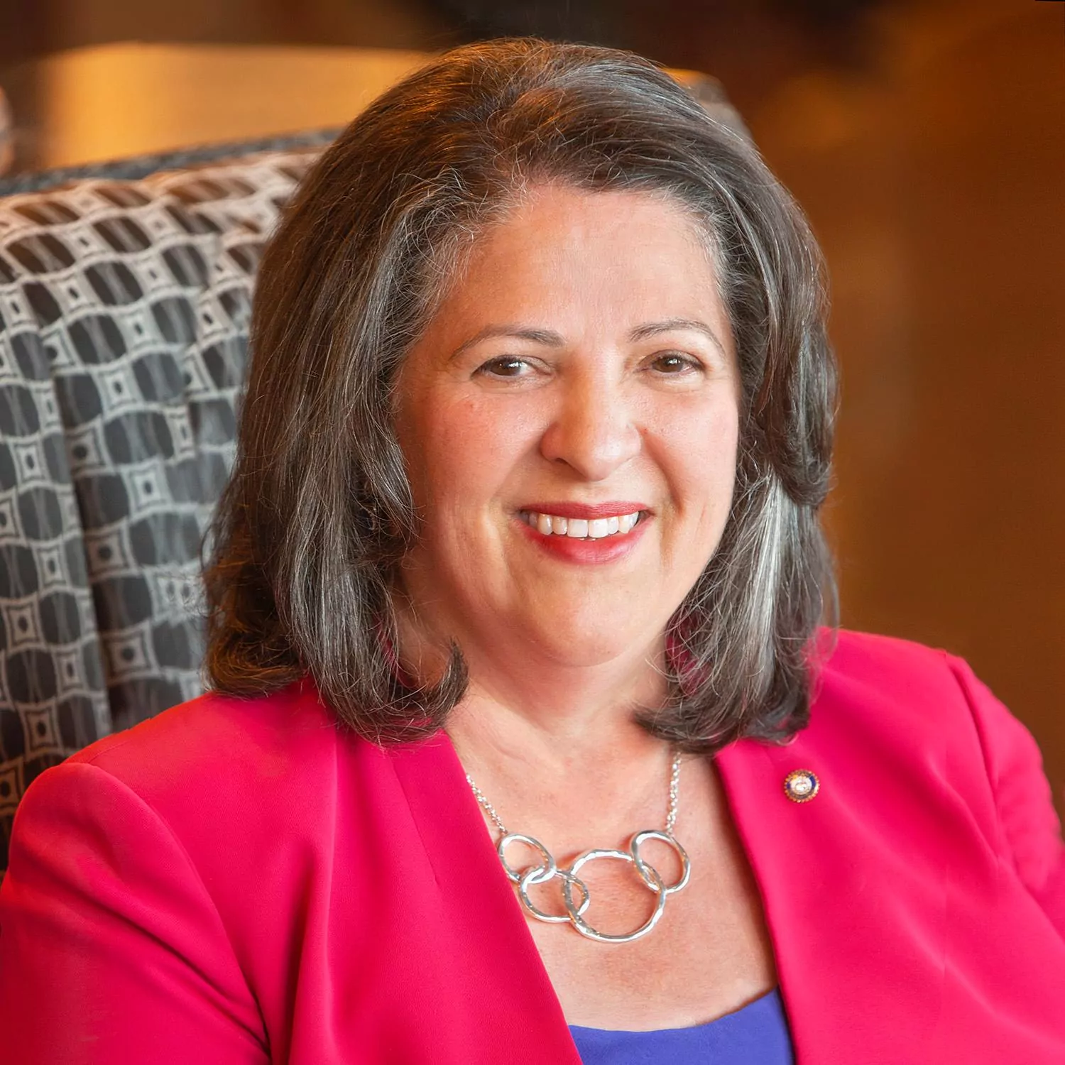 le forum du rotary isabelle martinez hayer, une femme aux cheveux gris longs, est assise sur une chaise à motifs, vêtue d'un blazer rose vif, d'un collier argenté et d'un haut bleu, et sourit à la caméra.