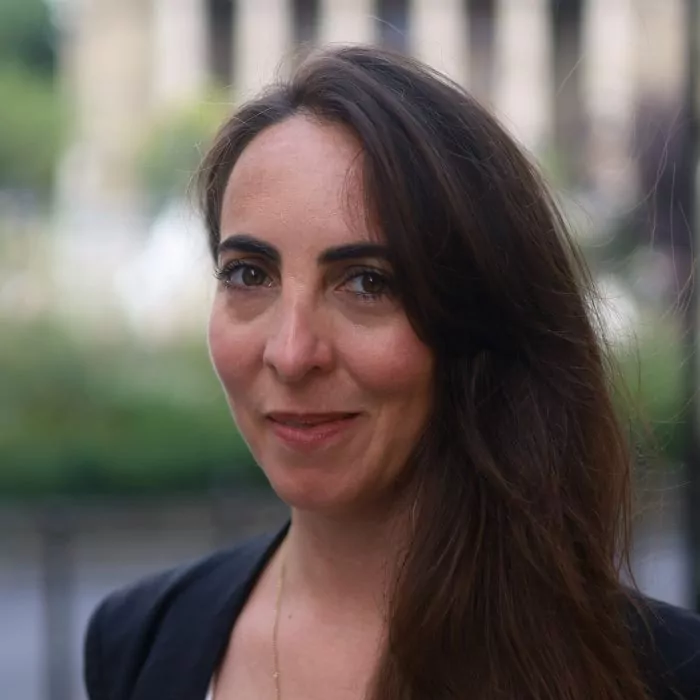 le forum du rotary une femme aux longs cheveux bruns et vêtue d'un blazer noir se tient à l'extérieur, regardant l'appareil photo avec un léger sourire. l'arrière plan est flou, montrant de la verdure et un bâtiment flou.