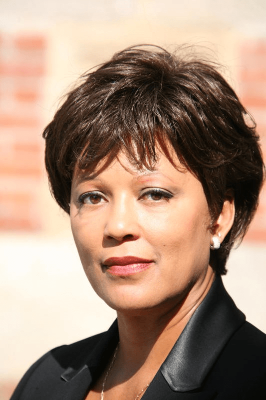 le forum du rotary une femme aux cheveux bruns courts, portant un blazer noir et des boucles d'oreilles en perles, regarde l'appareil photo avec une expression neutre. l'arrière plan est flou et laisse entrevoir un mur de briques.