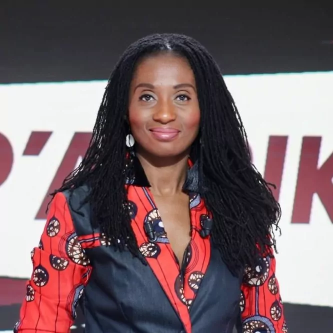 le forum du rotary une femme aux longs cheveux noirs tressés porte une chemise à motifs circulaires rouges, noirs et blancs. elle se tient devant un fond avec un texte partiel rouge et blanc. elle regarde la caméra et sourit légèrement.