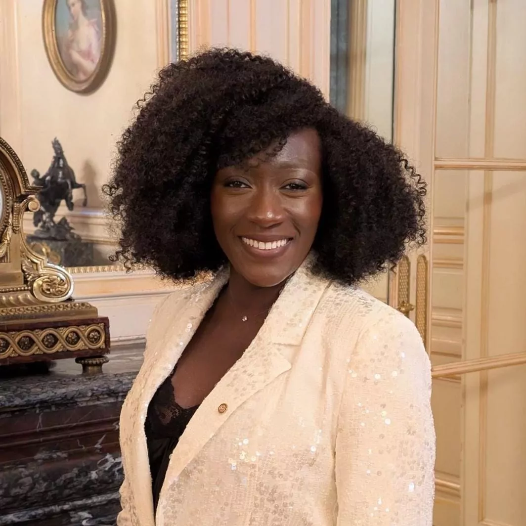 le forum du rotary une femme aux cheveux noirs et bouclés sourit à l'appareil photo. elle porte un blazer à paillettes de couleur crème sur un haut noir et se tient dans une pièce élégante avec du marbre, des touches d'or et un tableau encadré en arrière plan.