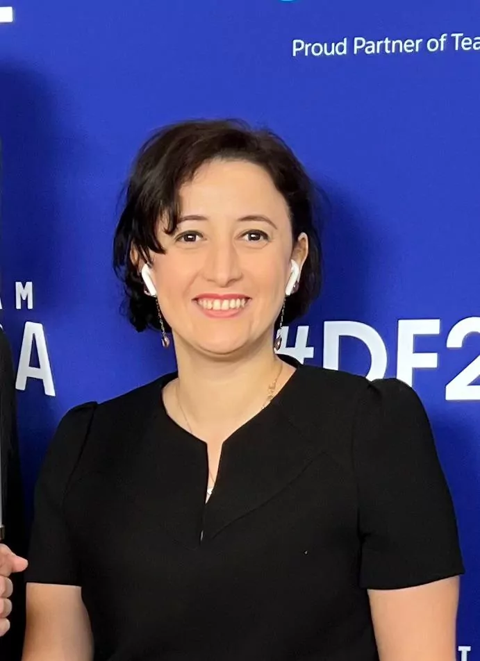 le forum du rotary fatiha zelmat, une femme aux cheveux bruns courts, portant un haut noir et des écouteurs sans fil blancs, sourit à la caméra devant un fond bleu avec du texte blanc et des hashtags.
