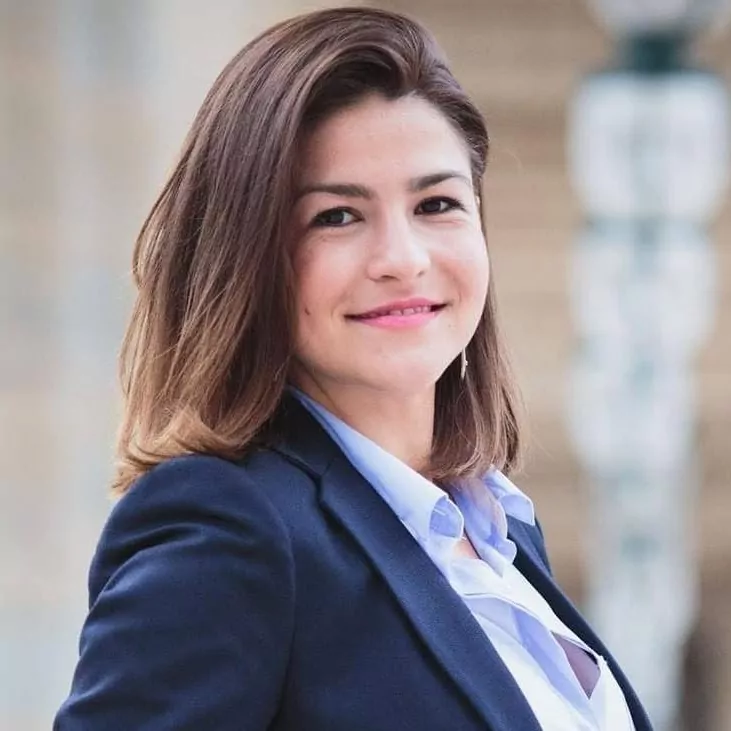 le forum du rotary une femme aux cheveux bruns mi longs, vêtue d'un blazer bleu marine et d'une chemise bleu clair, se tient à l'extérieur et sourit à l'appareil photo. l'arrière plan est flou.