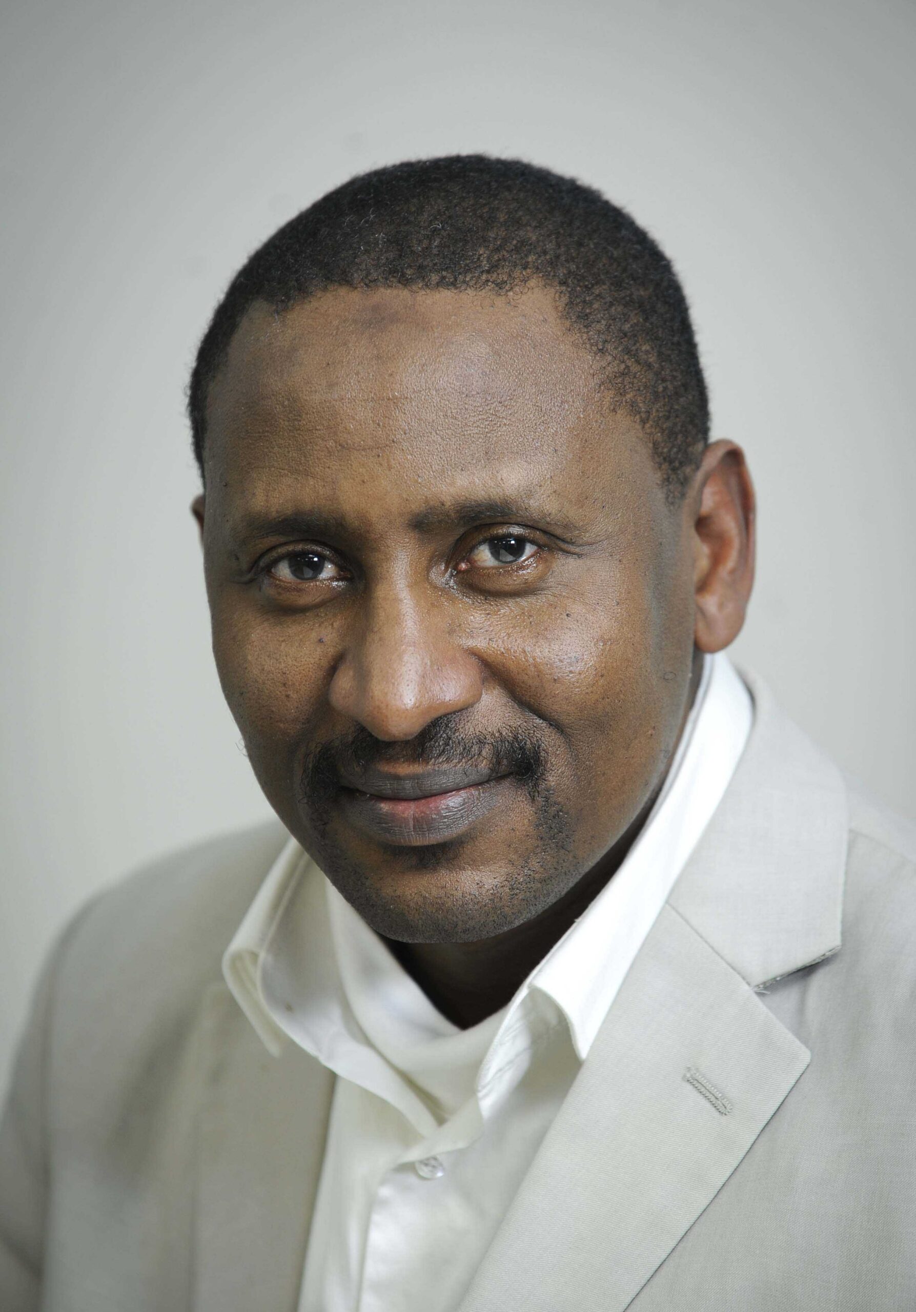 le forum du rotary un homme aux cheveux courts et à la moustache, vêtu d'un blazer de couleur claire et d'une chemise blanche, pose pour une photo de tête sur un fond clair et uni.