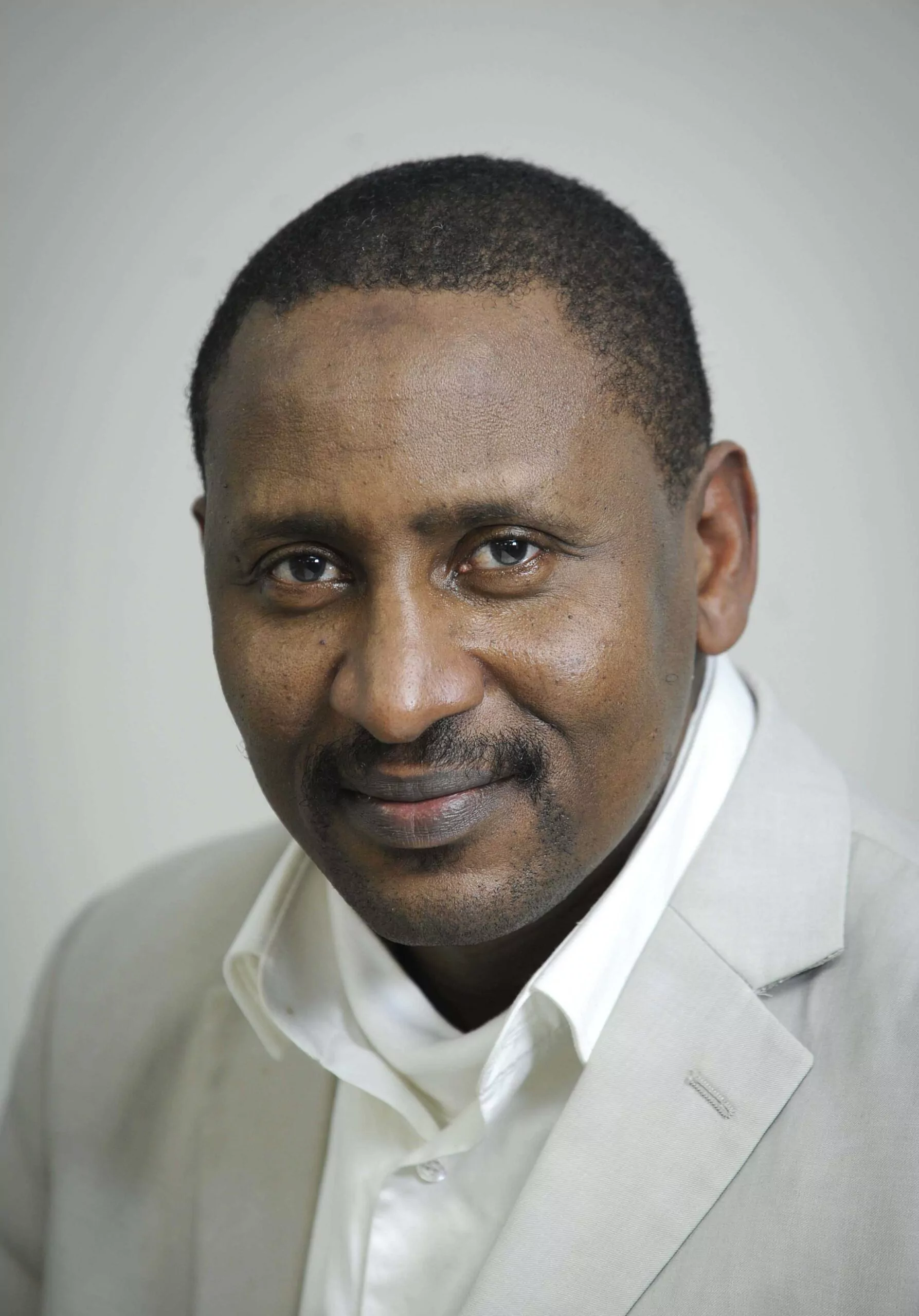 le forum du rotary un homme aux cheveux courts et à la moustache, vêtu d'un blazer de couleur claire et d'une chemise blanche, pose pour une photo de tête sur un fond clair et uni.