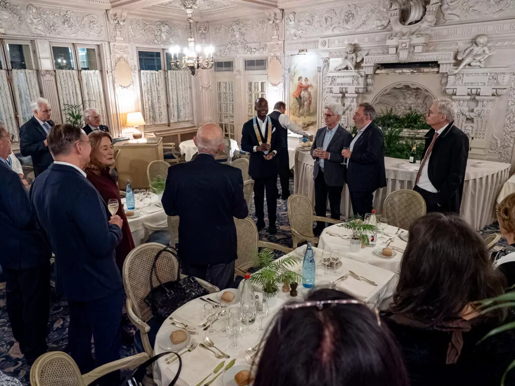 le forum du rotary un groupe de personnes en tenue de soirée se tient debout et discute dans une salle à manger ornée de murs décorés et d'une grande cheminée, tandis que certains tiennent des boissons et que d'autres s'assoient à des tables élégamment dressées.