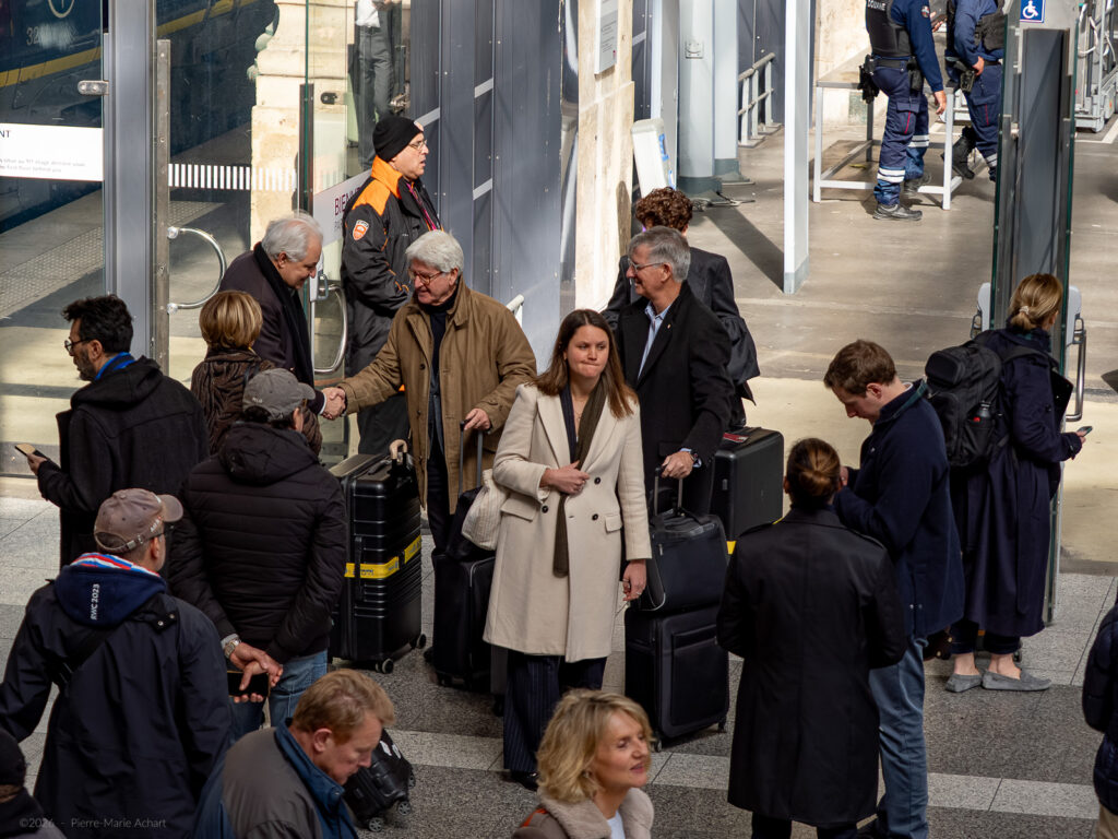 le forum du rotary un groupe de personnes se tient à l'extérieur d'un bâtiment avec des valises et des sacs. deux hommes se serrent la main, tandis que d'autres regardent leur téléphone ou parlent. des agents de sécurité sont visibles à l'arrière plan, près de l'entrée.