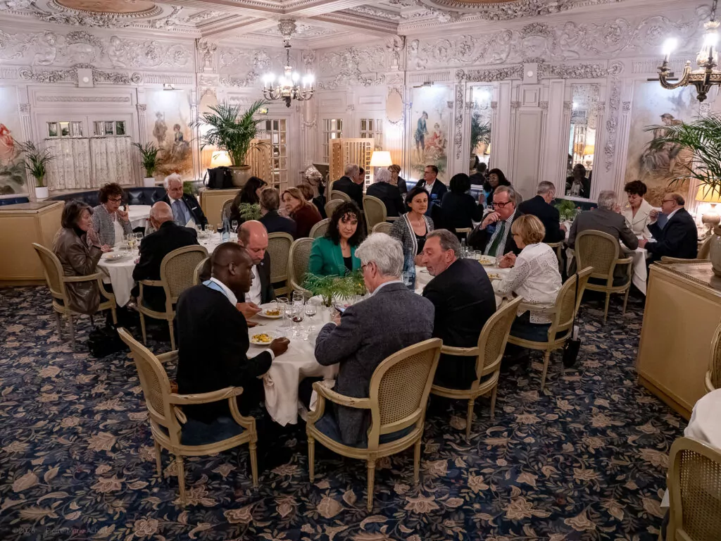le forum du rotary un groupe de personnes est assis à des tables rondes dans une salle à manger ornée et bien éclairée, aux murs blancs, aux moulures élaborées et à la moquette à motifs, et discute autour d'un repas.