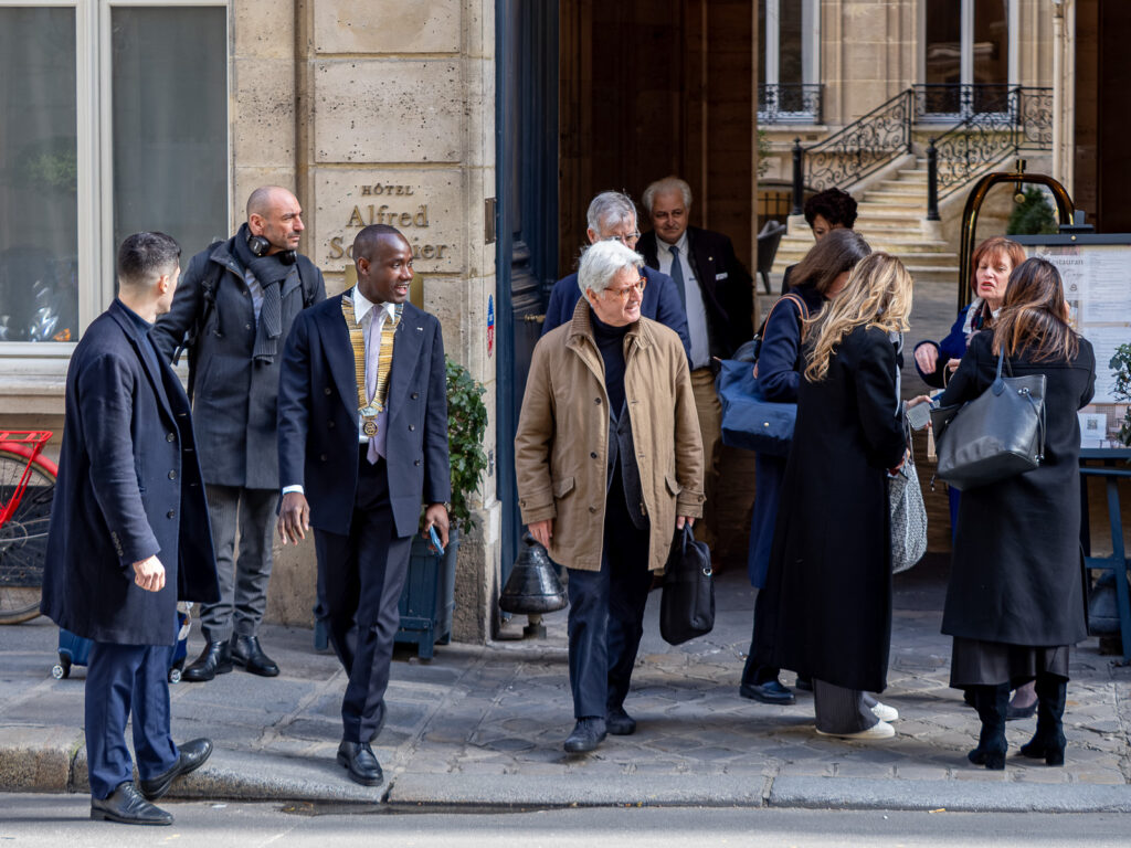 le forum du rotary un groupe de personnes vêtues de manteaux se tient debout et discute devant l'entrée de l'hôtel alfred sommier sur un trottoir de la ville. un homme porte un cordon et un autre tient un dossier. un vélo rouge est partiellement visible sur la gauche.