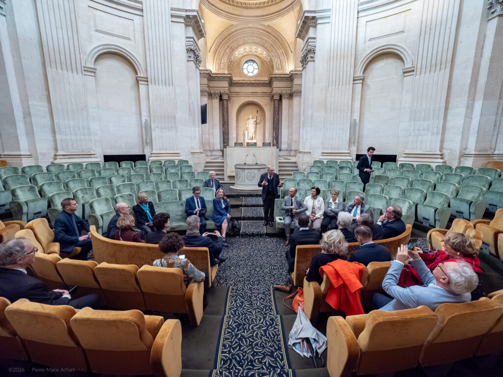le forum du rotary un groupe de personnes est assis en demi cercle dans une grande salle à l'architecture ornée, avec une statue dans une alcôve. une personne se tient debout et parle au centre, tandis que les autres écoutent. des rangées de sièges vides entourent le groupe.