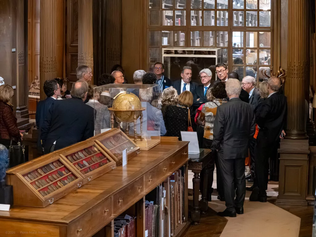 le forum du rotary un groupe de personnes, pour la plupart en tenue de travail, se tient dans une bibliothèque avec des étagères en bois, de vieux livres et un globe terrestre, écoutant quelqu'un qui parle au centre.