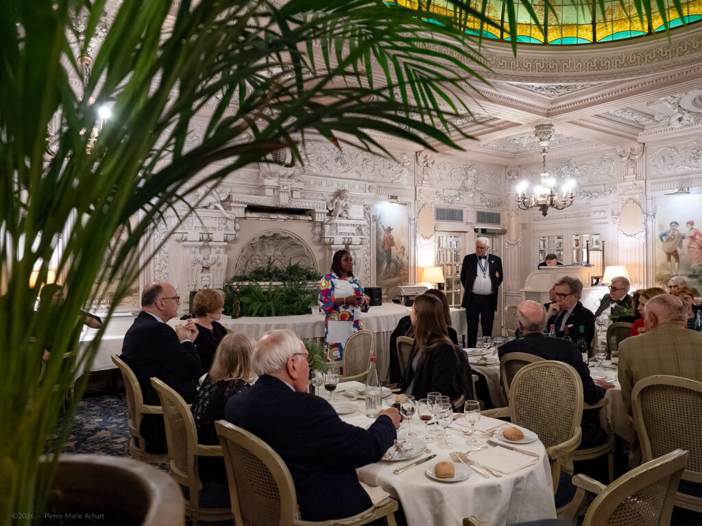 le forum du rotary un groupe de personnes est assis à des tables rondes dans une salle à manger ornée, écoutant une femme debout qui parle à l'avant. la pièce présente des sculptures murales blanches élaborées et un plafond en verre teinté.