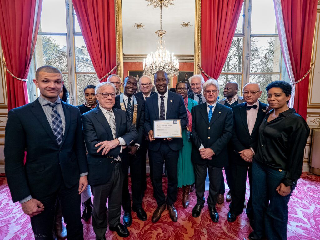 le forum du rotary un groupe de personnes habillées de manière formelle posant à l'intérieur, avec une personne au centre tenant un certificat encadré. elles se tiennent sur un tapis rouge dans une pièce ornée de rideaux rouges, de hautes fenêtres et d'un lustre.