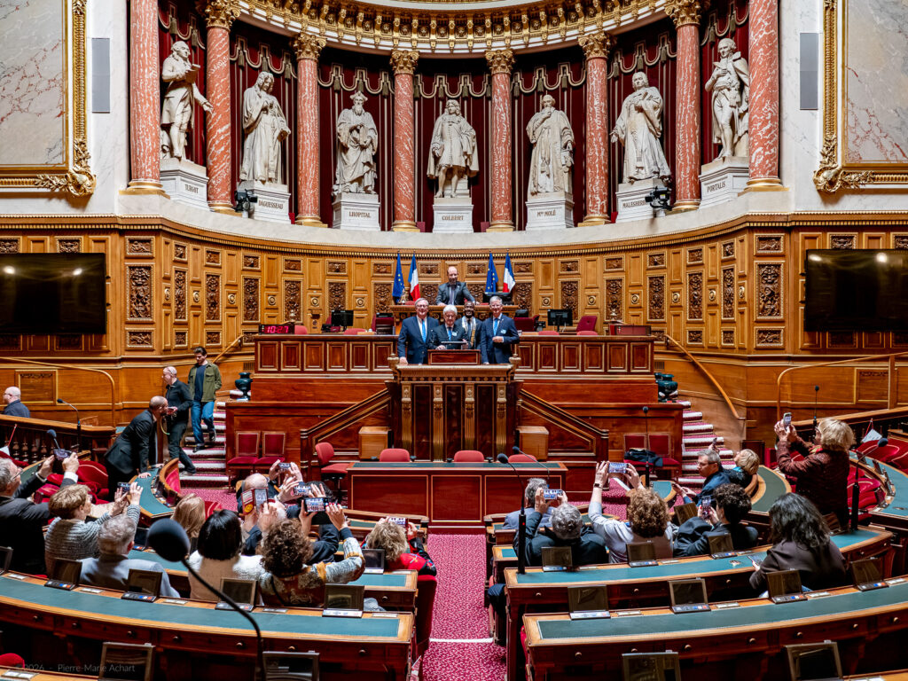 le forum du rotary un groupe de personnes se tient sur le podium central d'une grande salle législative avec des boiseries ornées, des statues et des sièges rouges. plusieurs drapeaux se trouvent derrière l'estrade et un public est assis devant.