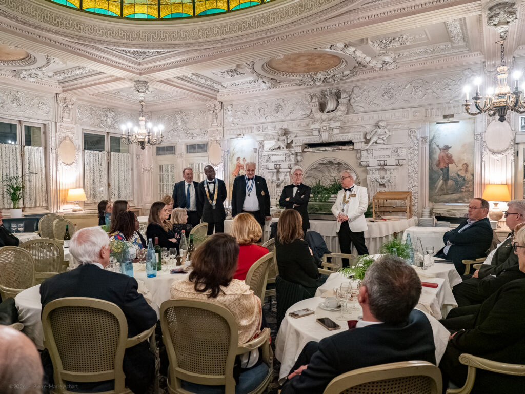 le forum du rotary un groupe de personnes est assis autour de tables dans une salle à manger ornée, tandis que six personnes se tiennent debout et parlent à l'avant. la pièce est ornée de plâtres élaborés, de lustres et d'une grande cheminée décorative.
