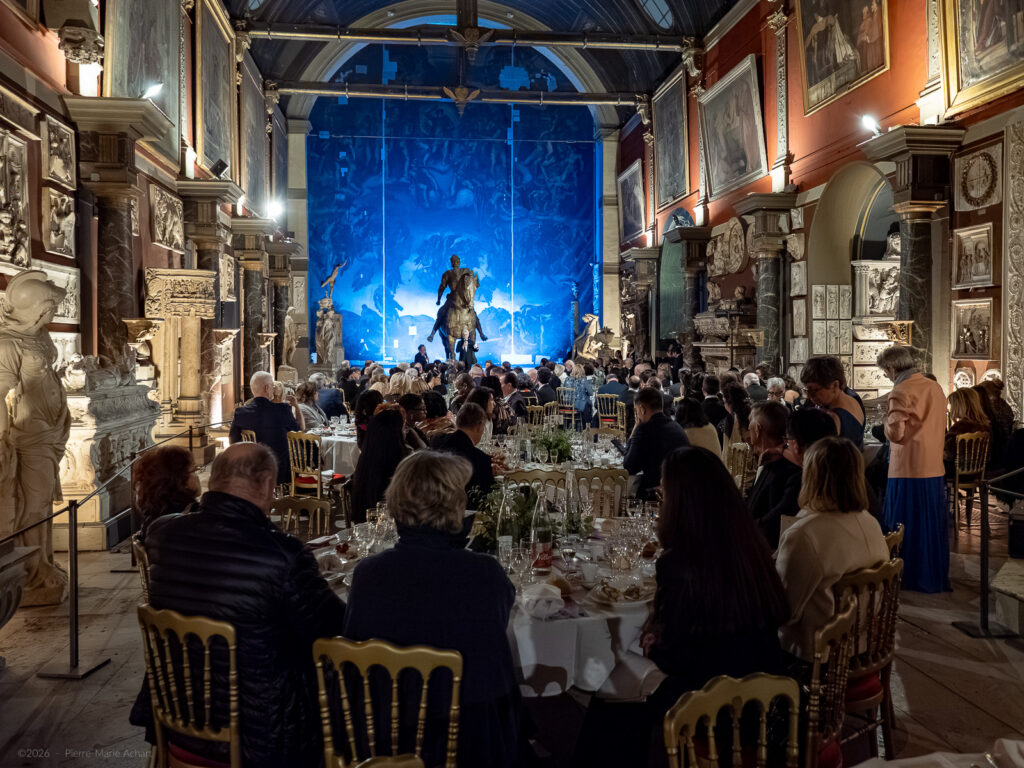 le forum du rotary un groupe de personnes assises à des tables dans une salle ornée de murs rouges et de sculptures classiques, regardant un spectacle sur une scène éclairée en bleu à l'avant de la salle.