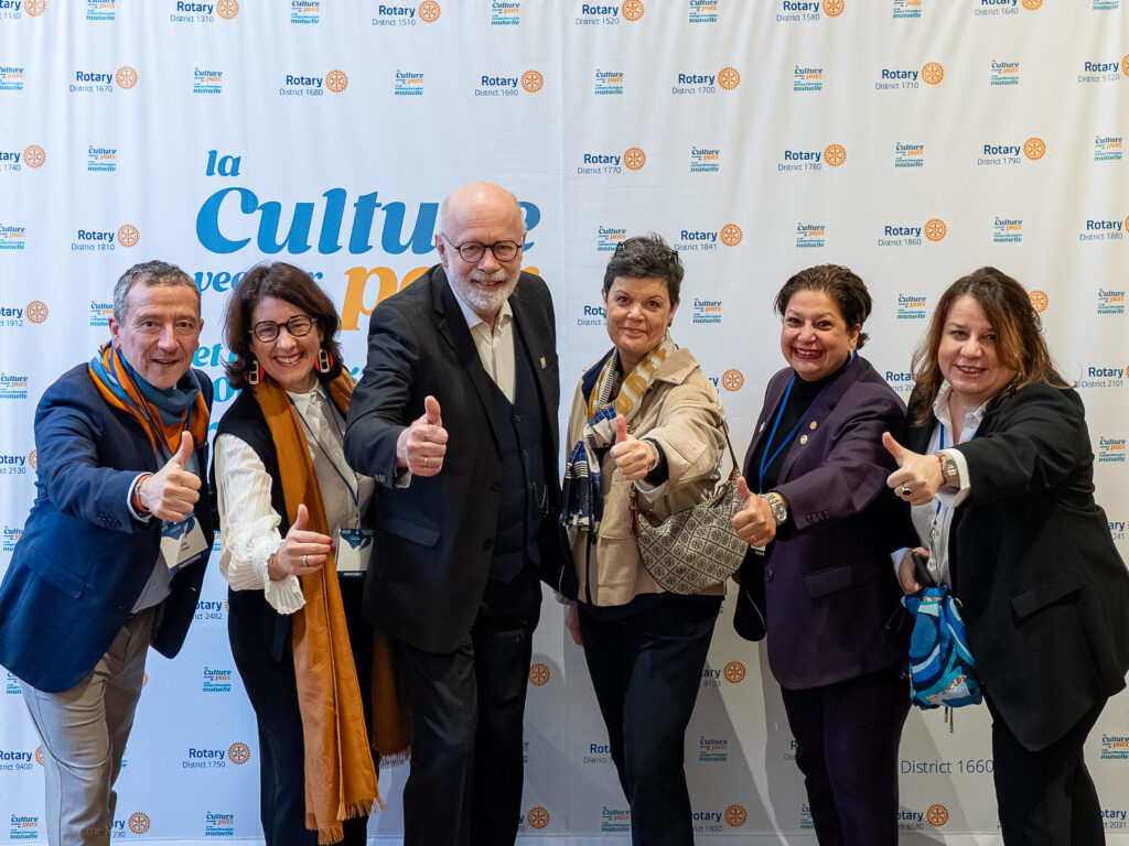 le forum du rotary six adultes se tiennent devant une bannière du rotary, souriant et levant le pouce. ils sont vêtus de tenues professionnelles ou semi formelles et portent des badges ou des écharpes. l'arrière plan présente les logos du rotary et de la manifestation.