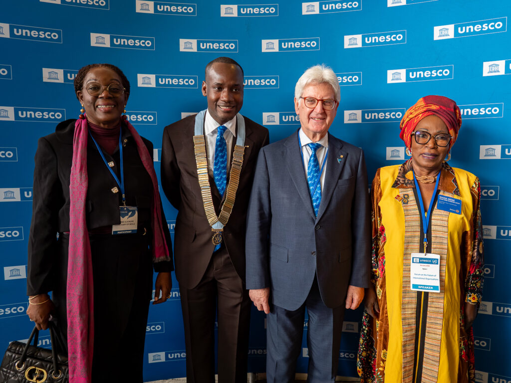 le forum du rotary quatre personnes se tiennent côte à côte devant une toile de fond bleue de l'unesco. elles sont habillées de manière formelle et portent des badges visibles. un homme porte une chaîne de cérémonie autour du cou. ils posent pour une photo de groupe.
