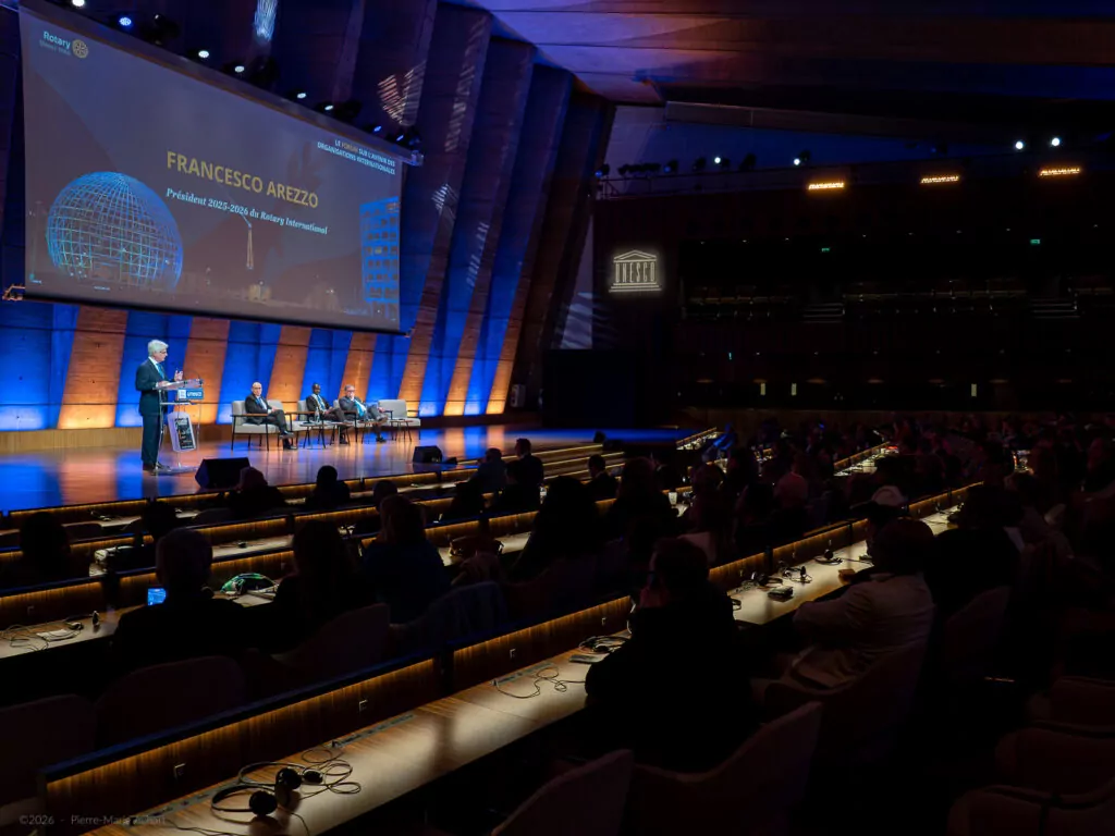 le forum du rotary un orateur se tient sur un podium, sur une scène avec un grand écran affichant "francesco arezzo" et un public assis dans un auditorium moderne faiblement éclairé avec une lumière bleue.