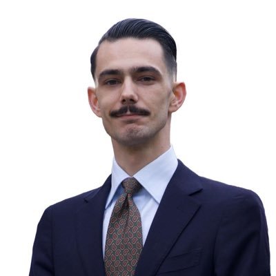 le forum du rotary un homme aux cheveux noirs gominés et à la moustache, portant un costume sombre, une chemise blanche et une cravate à motifs, pose sur un fond blanc uni.