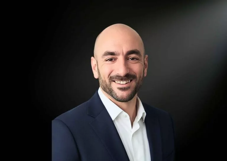le forum du rotary un homme au crâne rasé, à la barbe et à la moustache taillées, vêtu d'une veste de costume sombre et d'une chemise blanche, sourit à l'appareil photo sur un fond sombre et légèrement éclairé.