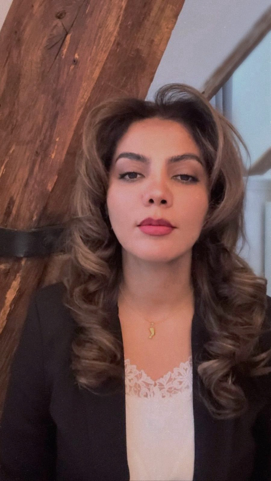 le forum du rotary une femme aux longs cheveux bruns ondulés porte un blazer noir sur un haut en dentelle blanche. elle se tient devant une poutre en bois et regarde directement la caméra avec une expression neutre.