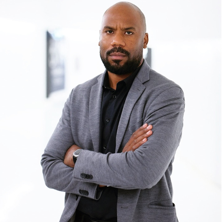 le forum du rotary un homme barbu et rasé se tient debout, les bras croisés, vêtu d'un blazer gris sur une chemise noire. il regarde directement la caméra avec une expression neutre sur un fond blanc et flou.